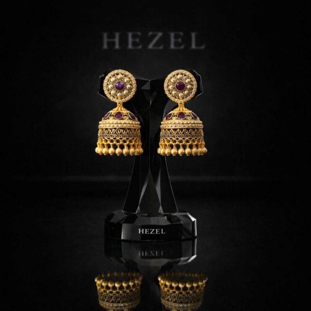 Elegant gold jhumka earrings with intricate detailing and ruby accents by Hezel, displayed on a premium black stand with a luxury backdrop.