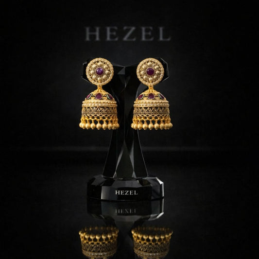 Elegant gold jhumka earrings with intricate detailing and ruby accents by Hezel, displayed on a premium black stand with a luxury backdrop.