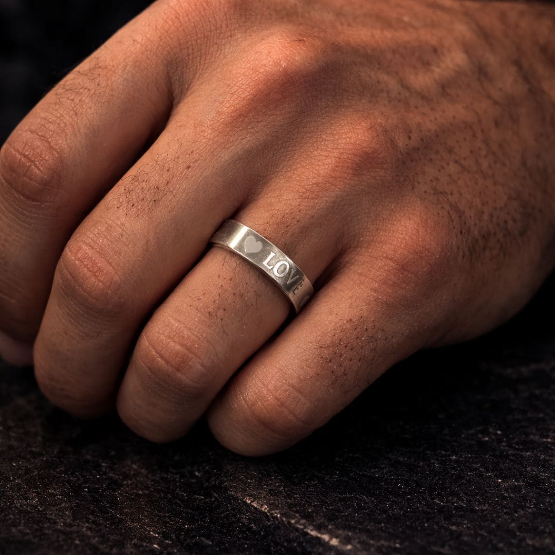 Minimalist silver band ring engraved with “LOVE” and a heart, worn on a hand against a dark textured surface