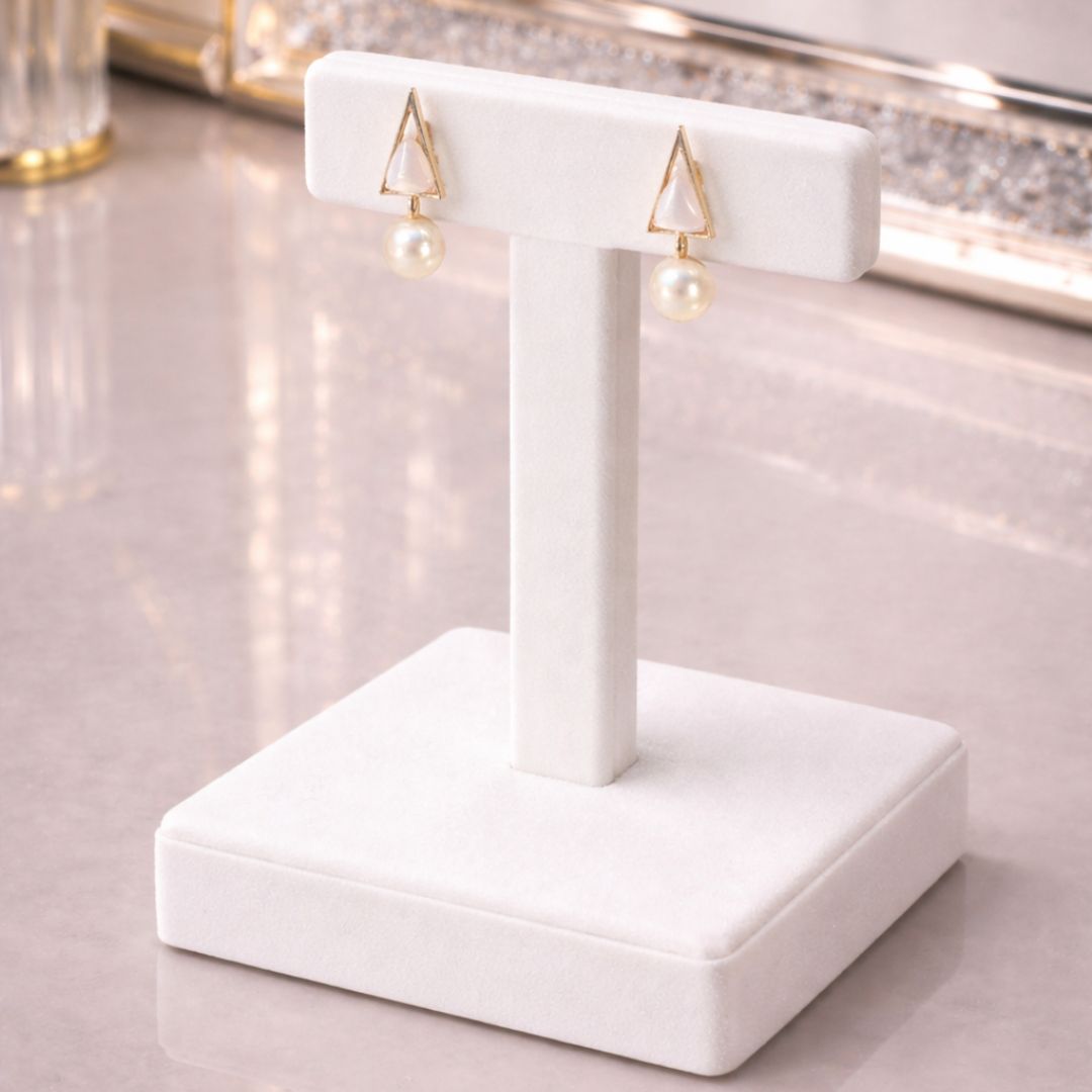 Hezel gold triangle stud earrings with pearl drops, displayed on a white stand in a soft elegant setting.